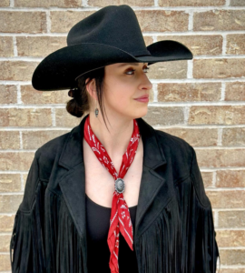 Taylor Teasley wearing a black cowboy hat and fringed black jacket with a red bandana, standing in front of a brick wall.