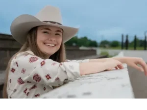 Tori Hughes smiling in a white cowboy hat and patterned shirt while leaning against a white railing outdoors.