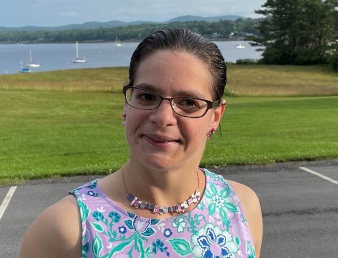Emilia Sandona smiles while standing outdoors beside a black railing, with a scenic waterfront, sailboats, and green landscape in the background. She is wearing a purple floral sleeveless dress, glasses, and colorful jewelry.